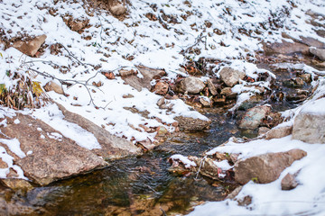 Meltwater stream running through the stones and surrounded by brown melting snow and dry grass at mountains.