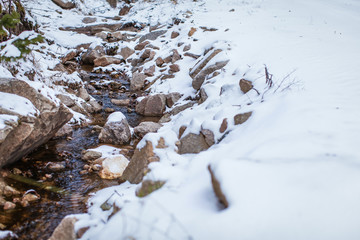 Meltwater stream running through the stones and surrounded by brown melting snow and dry grass at mountains.