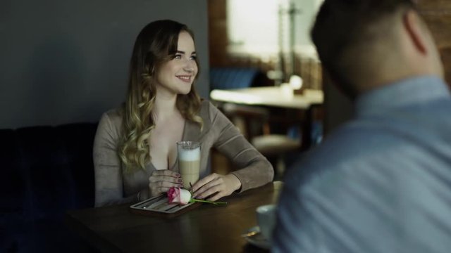 Beautiful blonde with wide beautiful smile and big breast/boobs in tight dress with decolette sitting in a cafe, posing to a camera phone of her man. There are a cocktail and a rose on a table