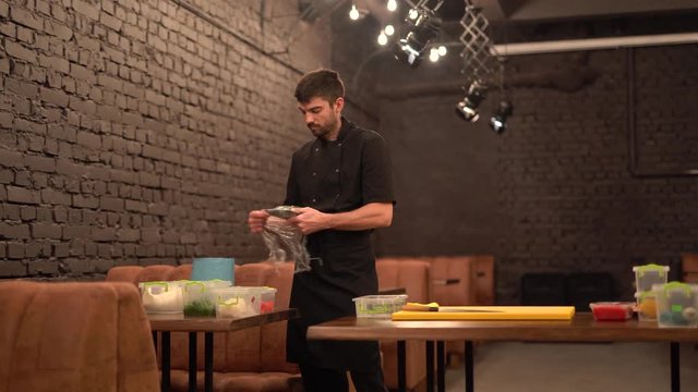 Hansome male waiter with black bear and moustache wearing black apron/uniform unpacking products in basement, tearing off food film covering lunchboxes to get vegetables out. Working atmosphere