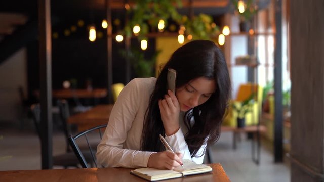 Very beautiful girl with black long hair in white blouse sitting alone in a cafe while writing sth down and talking on the phone. Pretty brunette is dived into her notebook like business lady