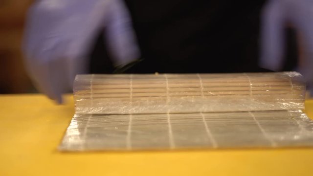 Male hands in blue rubber gloves with visible black torso making sushi with the cheese outside, rolling them in bamboo, splashing sushi and dividing them. Closeup concept. Chef in restarant