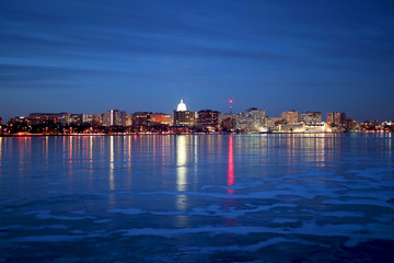 Madison downtown skyline illuminated at winter night with official buildings, Monona Terrace and capitol dome, glowing in the dark. Cityscape reflects in a frozen lake Monona.