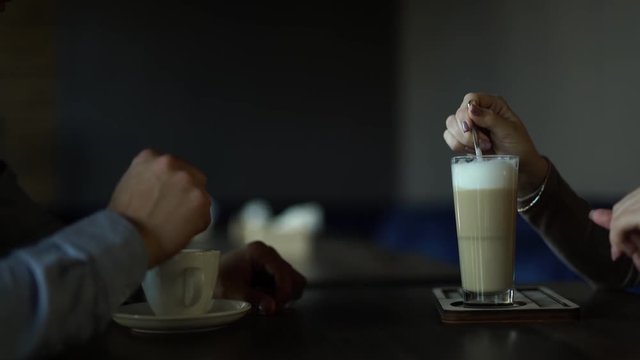 Couple/lovers sitting opposite each other in a cozy cafe with hands profile. Their hands are stirring/mixing their coffe/cocktail/cappucino/latte with the dessert spoon. Love athmosphere