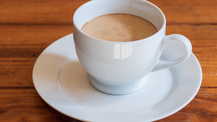Cup of coffee with cream in white ceramic cup, on small white ceramic plate, on wooden surface