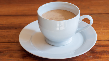 Cup of coffee with cream in white ceramic cup, on small white ceramic plate, on wooden surface