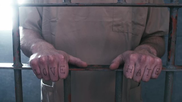 Close-up dolly shot of a prisoner standing at the door of his cell, holding the bars. You can see that he has "love" and "hate" tattooed on his fingers and then he walks away.