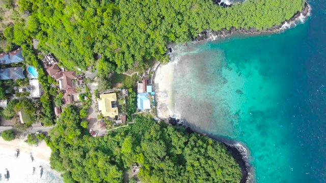 Amazing aerial view taken by a flying drone, slow upward rise to a remarkable height, of a tourist resort by the sea in Padang Bai, Bali. Small bungalows, trees and a sandy beach.