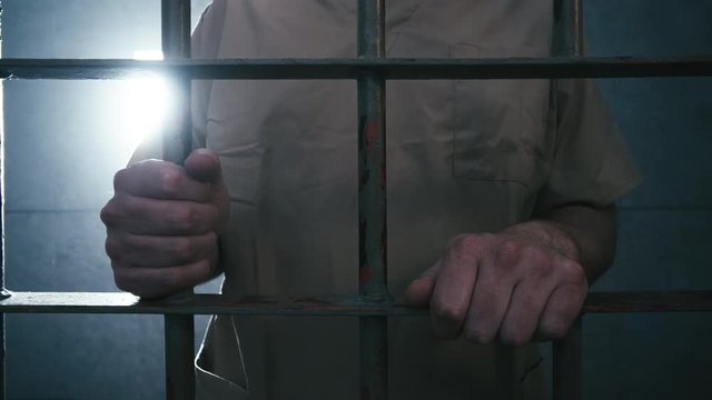 Close-up tracking shot from left to right of a prisoner holding onto the bars of his cell. When the camera stops moving he walks off screen.