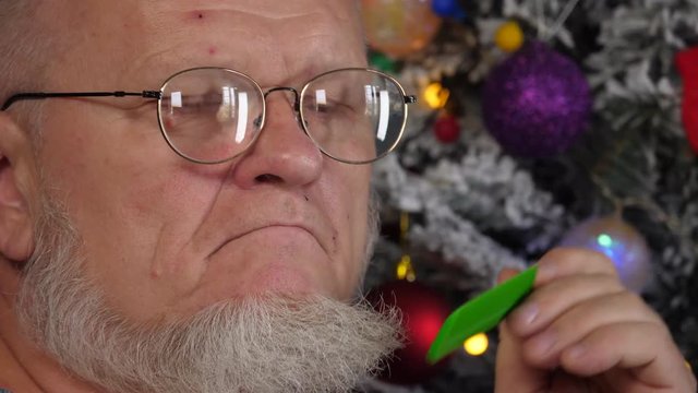 Happy elderly man combing his gray beard with comb on background of Christmas tree in garlands, green UFO balls, proton purple toy, plastic pink angel, neon glow