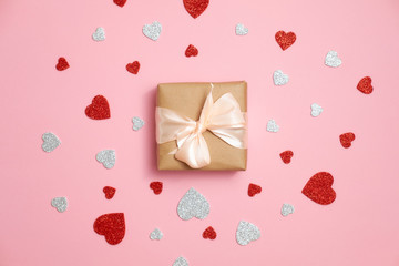 Valentines day composition : gift box packed in kraft paper with ribbon bow and red and grey heart shaped valentines card lay on pink background woman's desk. Surprise for lovers, love concept.