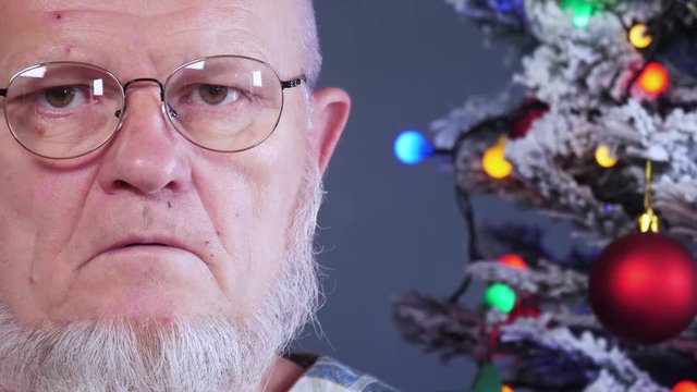 happy elderly man grimaces and actively expresses his emotions against background of Christmas tree in garlands, green UFO balls, proton purple toy, plastic pink angel, neon glow