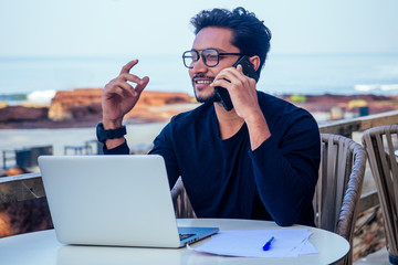 man talking on the phone india summer holidays.stylish young indian male freelancer working with laptop freelance surfing online.businessman at remote work on the beach.dream job.online purchase sale