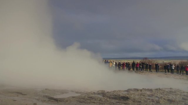 Tourists watch a spectacular eruption of The Great Geysir, one of Icelands best geysers. Its a sunny day with a row of people watching in the background. They are taking photos and having fun.