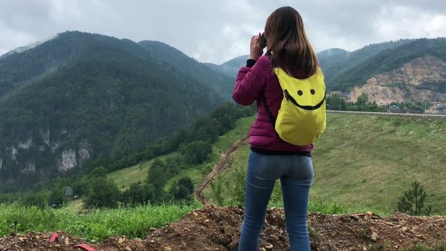 Woman traveler takes photos in scenic place in nature in cloudy rainy day from high poin. Back view