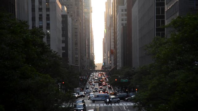 Looking down a busy New York City Street as vehicles try to get around under a beautiful sunset seen through the canyon of skyscrapers.