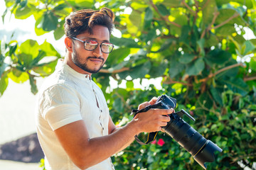 handsome and confident indian man photographer with a large professional camera taking pictures photo shooting on the beach.photo session on summer holiday on the background of green tropical trees