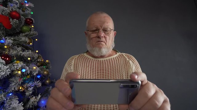 Happy Elderly man in Santa Claus hat is chatting with friends using smartphone and actively expresses his emotions against background of Christmas tree in garlands, green UFO balls, proton purple toy