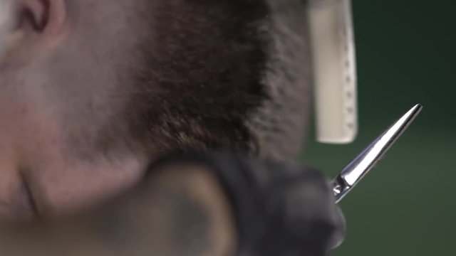 Tattooed barberman's hands in black gloves cutting customer brown hair using scissors and a comb for making a new style. Vertical format. Close-up concept