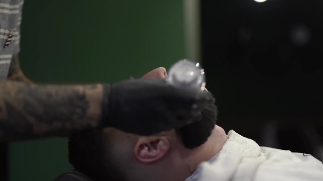 The customer lying on a special pillow in a barbershop. Barberman sweeping away/cleaning down the hair off client's face with the big brush. Close-up concept