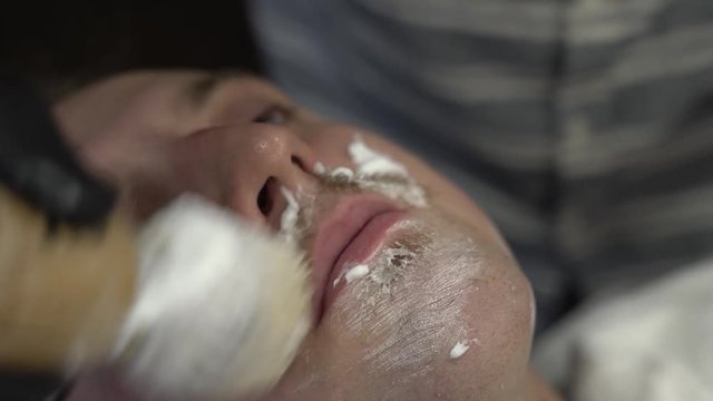 Barberman in black gloves applying shaving cream/gel on man's face with big brush. Client with eyes open. Close-up concept