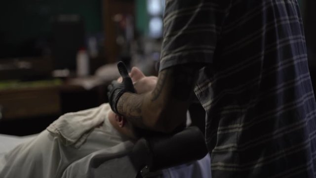 Barberman in black gloves spaying and spreading/rubbing special green liquid/cream over the customer's face in barbershop. Client's lying and relaxing under a white peignoir