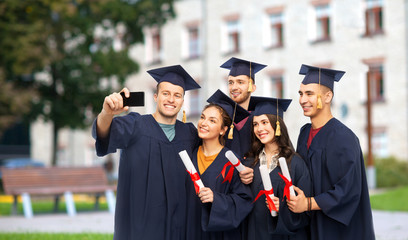 education, graduation and people concept - group of happy graduate students in mortar boards and bachelor gowns with diplomas taking selfie by smartphone over campus building background