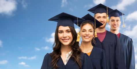 education, graduation and people concept - group of happy graduate students in mortar boards and bachelor gowns over blue sky and clouds background