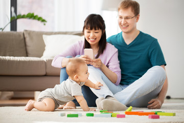 Fototapeta na wymiar mixed race family, parenthood and people concept - happy mother with smartphone, father and baby boy playing toy blocks at home