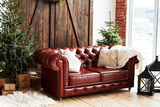 Leather vintage brown chesterfield sofa in Christmas interior with fir tree and gift boxes in loft room with wooden door and panoramic window, copy space