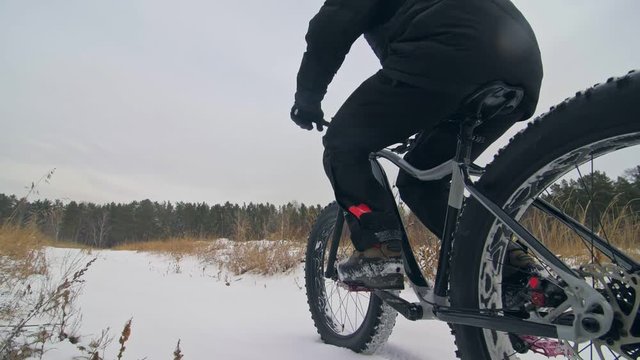 Professional extreme sportsman biker riding fat bike in outdoors. Cyclist ride in winter in snow field, forest. Man does trick on mountain bicycle with big tire in helmet, glasses. Slow motion 180fps.