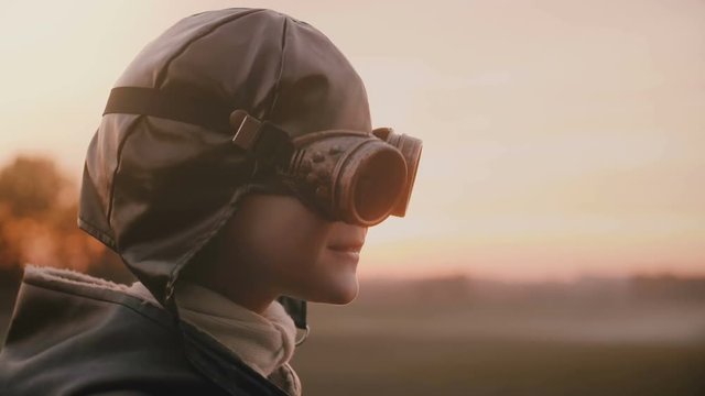 Little boy in old pilot costume putting on retro aviation glasses smiling in sunset field close-up slow motion.