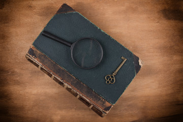 Old book, a key and a magnifying glass on the book against a worn wooden background with dark edges...