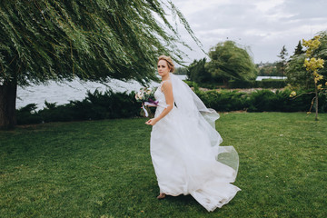 Blonde bride walks through the park with green grass and willows. Wedding portrait of a beautiful bride with a long dress. Wedding photography.