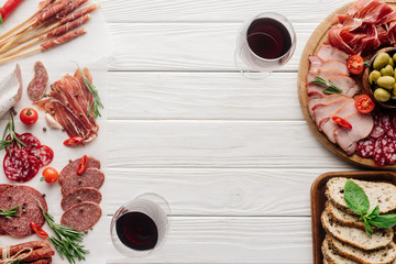 top view of glasses of red wine and delicious meat snacks on white wooden tabletop