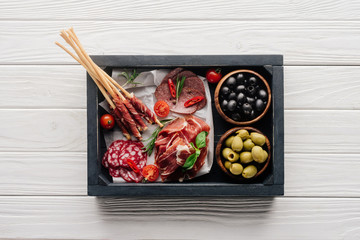 top view of meat appetizers, basil leaves, rosemary and olives in wooden box on white wooden backdrop