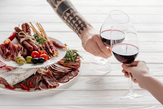 partial view of couple clinking glasses with red wine at white tabletop with meat appetizers - Powered by Adobe