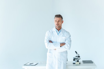 handsome scientist standing with crossed arms near table isolated on white