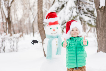 Smiling boy in red christmas hat with snowman on background showing thumbs up. Empty space for text