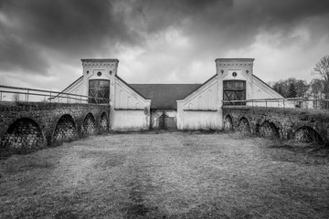 Vintage rural scene, dramatic front view of two black and white old rustic worn stone barn building farmhouse with dark sky. Two arched stone bridges in foreground.