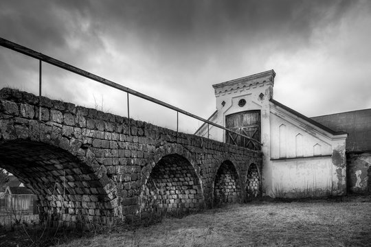 Vintage rural scene, dramatic front view of black and white old rustic worn stone barn farmhouse building with dark sky. Arched stone bridge in foreground.