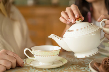 Hands of young woman with white china teapot pouring tea into cup of her senior mother