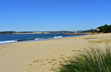 Beach with lighthouse, vegetation in sand dunes and small waves with foam. Golden sand, blue sea, clear sky. Sunny day, Galicia, Spain.