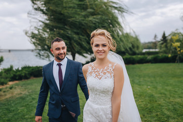 Blond bride and bearded groom walk, holding hands, in the park with green grass and willows. Wedding portrait of beautiful newlyweds. Wedding photography.