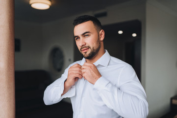 Stylish groom with a beard puts a shirt on. Morning groom. Wedding interior. Fees groom in the room.
