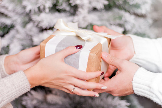 Closeup view of hands with gift box. Giving and receiving present on Christmas event. Love and romance couple celebrate xmas or New year holiday. X-mas fir tree on background.