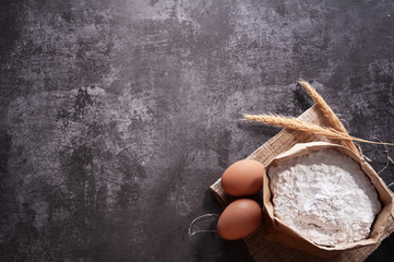 Bread ingredients with wheat ears, eggs and bag of flour on gray background. Top view