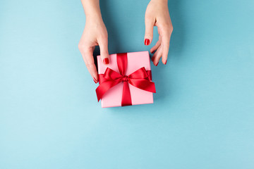 Overhead view on female hands with pink gift box wrapped with red bow on blue background. Minimal styled composition.