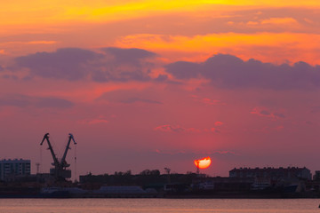 A huge red sun disk sets in the orange clouds beyond the city skyline into the silhouettes of houses and the sea loading dock. Coastal river sunset, Astrakhan, Russia.