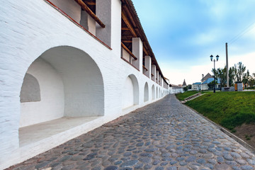 White stone wall of the courtyard with large arches and a wooden roof along the road with masonry...